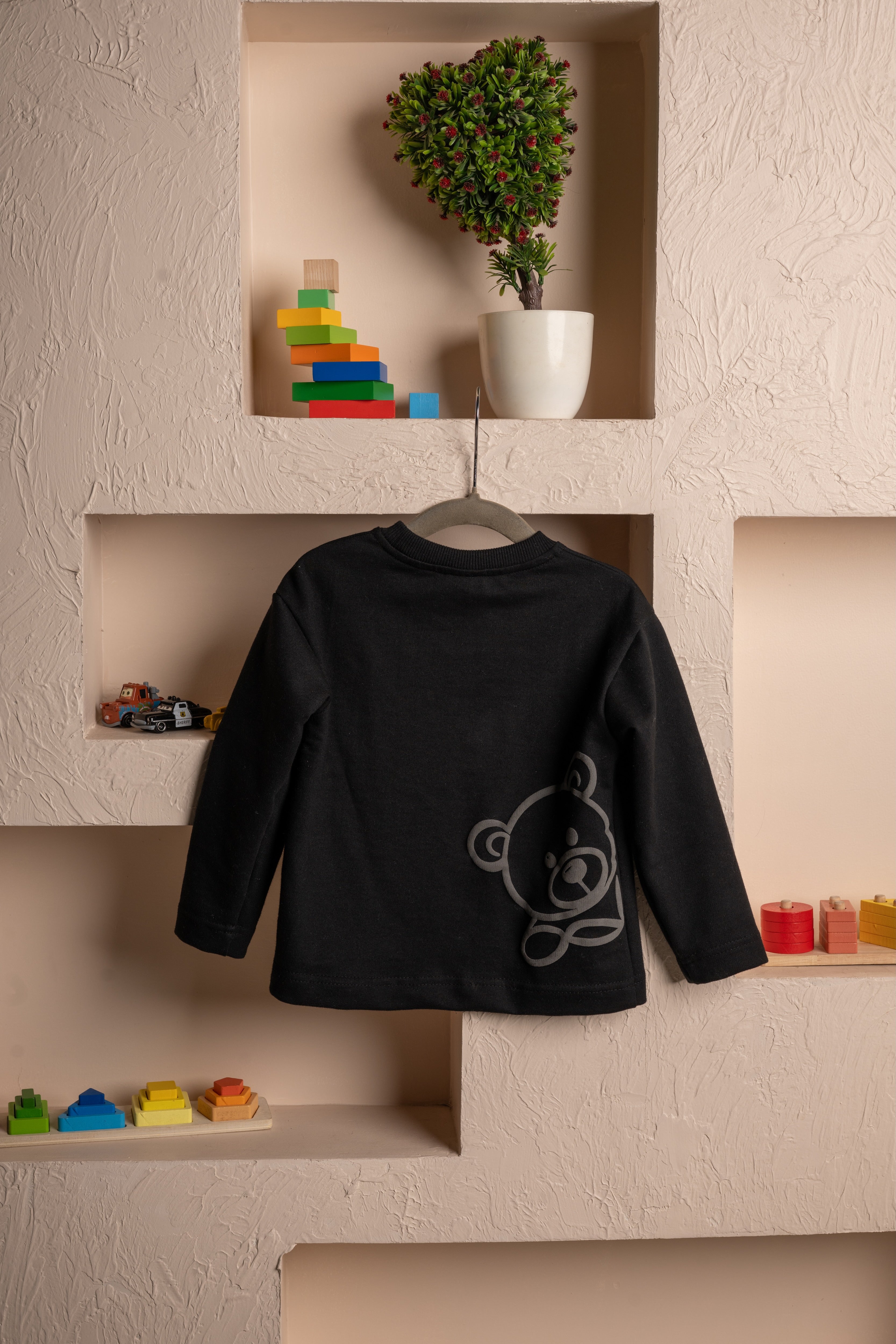 Luxurious black children's sweatshirt with teddy bear print hanging on textured beige wall shelves with colorful wooden toys and potted plant