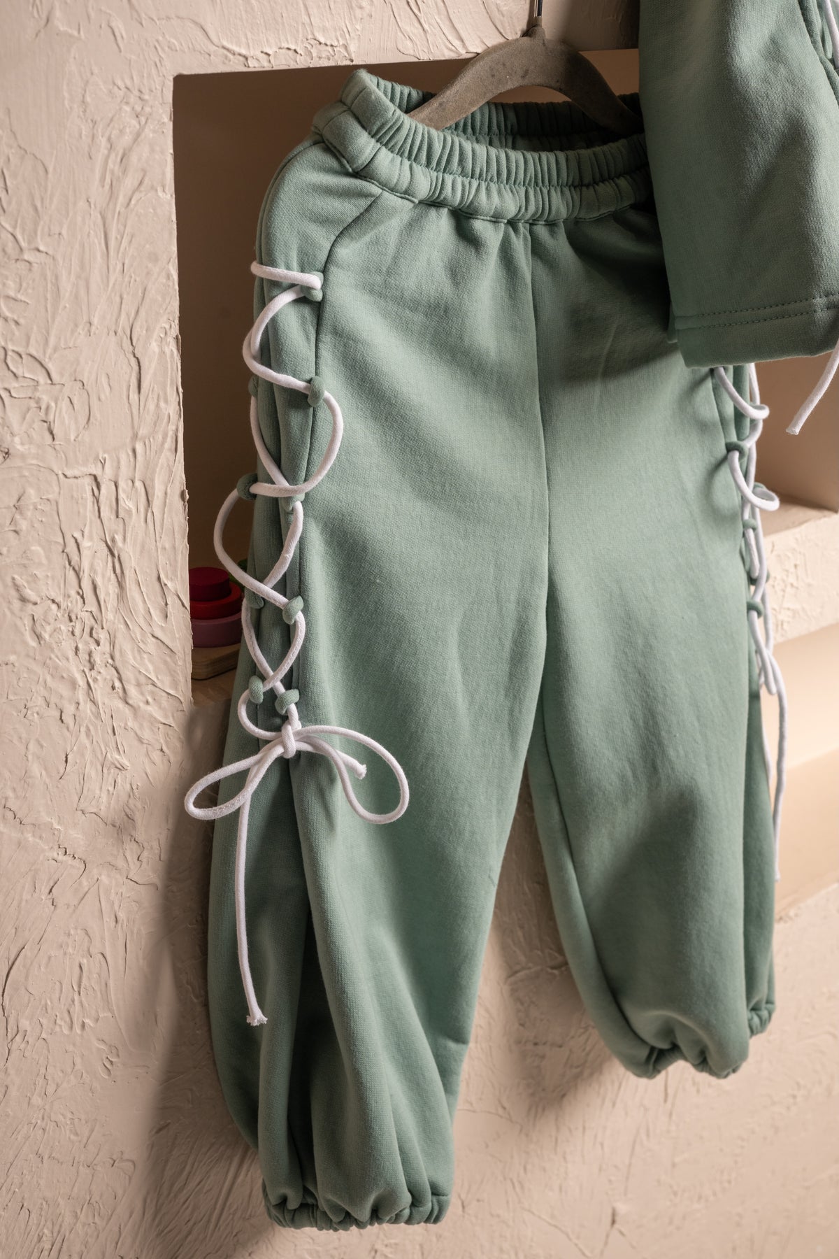 Elegant sage green joggers with white lace-up sides, hanging on textured beige wall