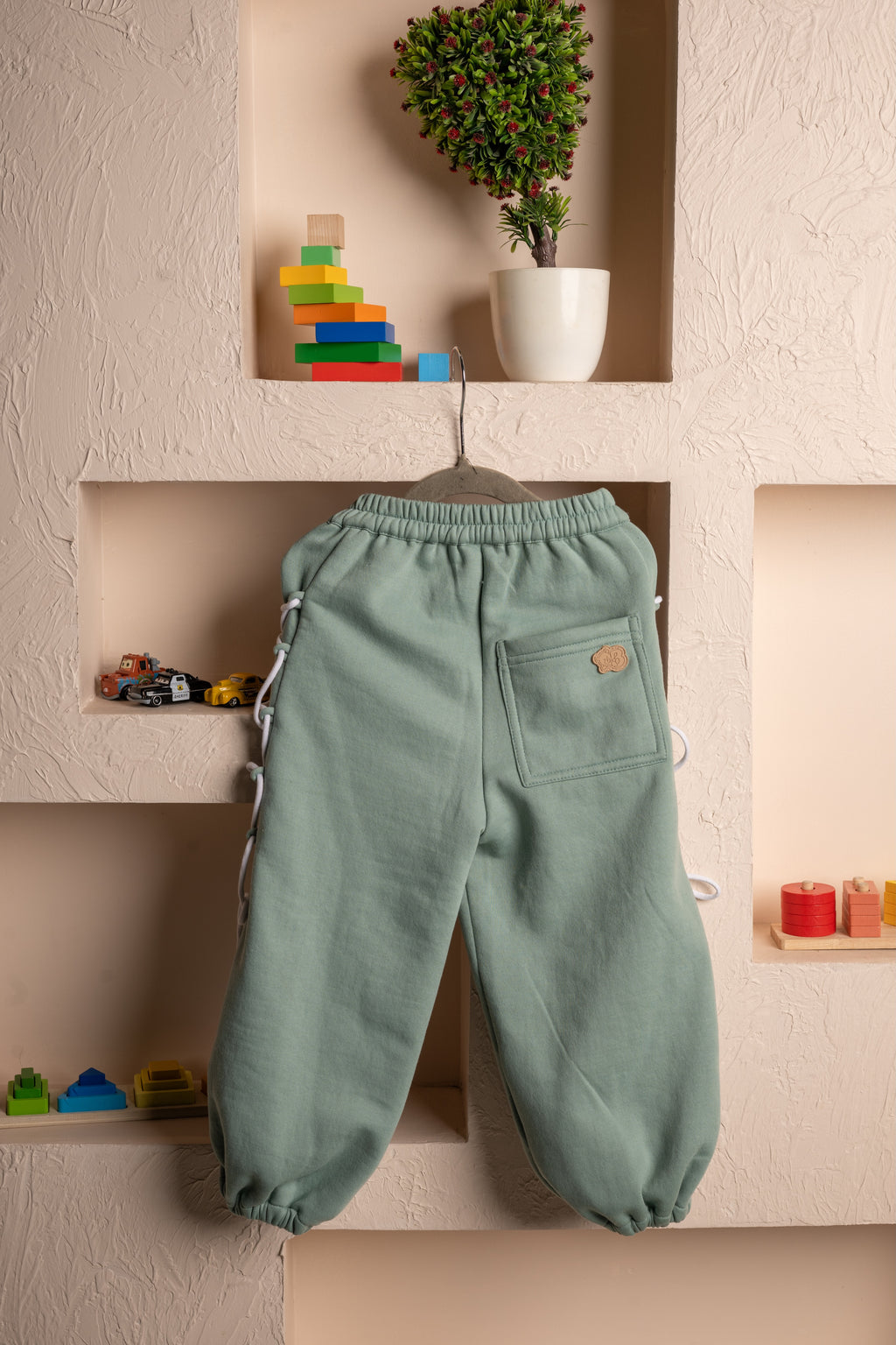 Elegant kids sage green sweatpants with side lace-up details, displayed on textured beige wall with colorful toys
