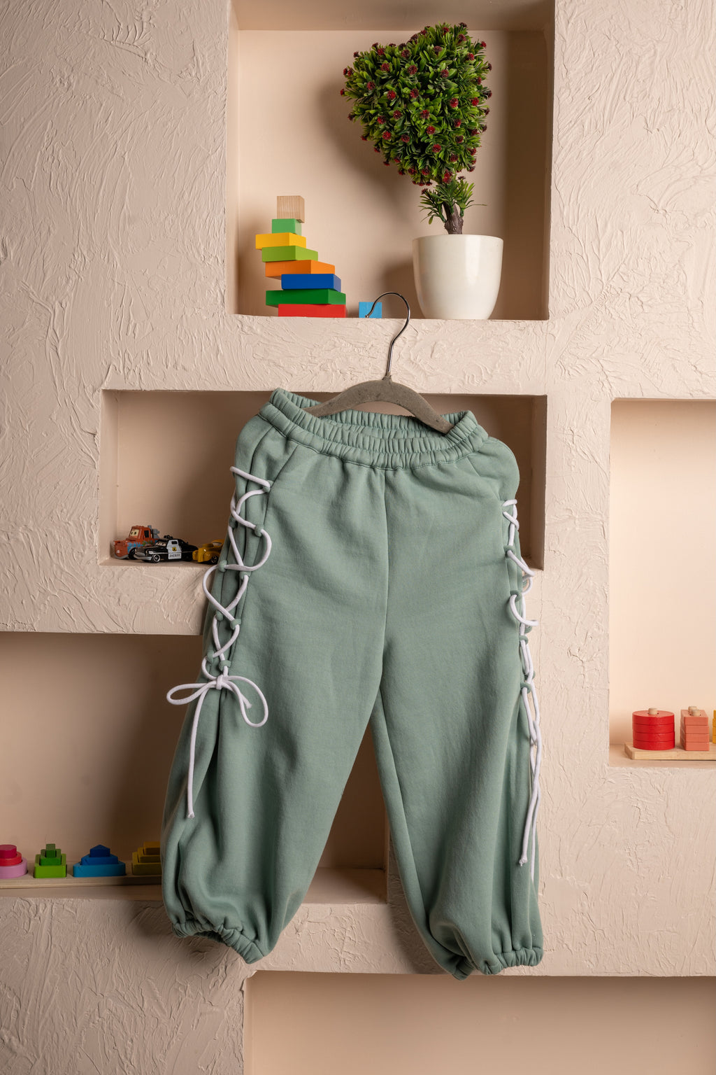 Elegant sage green jogger pants with white lace-up sides displayed on a textured beige wall niche