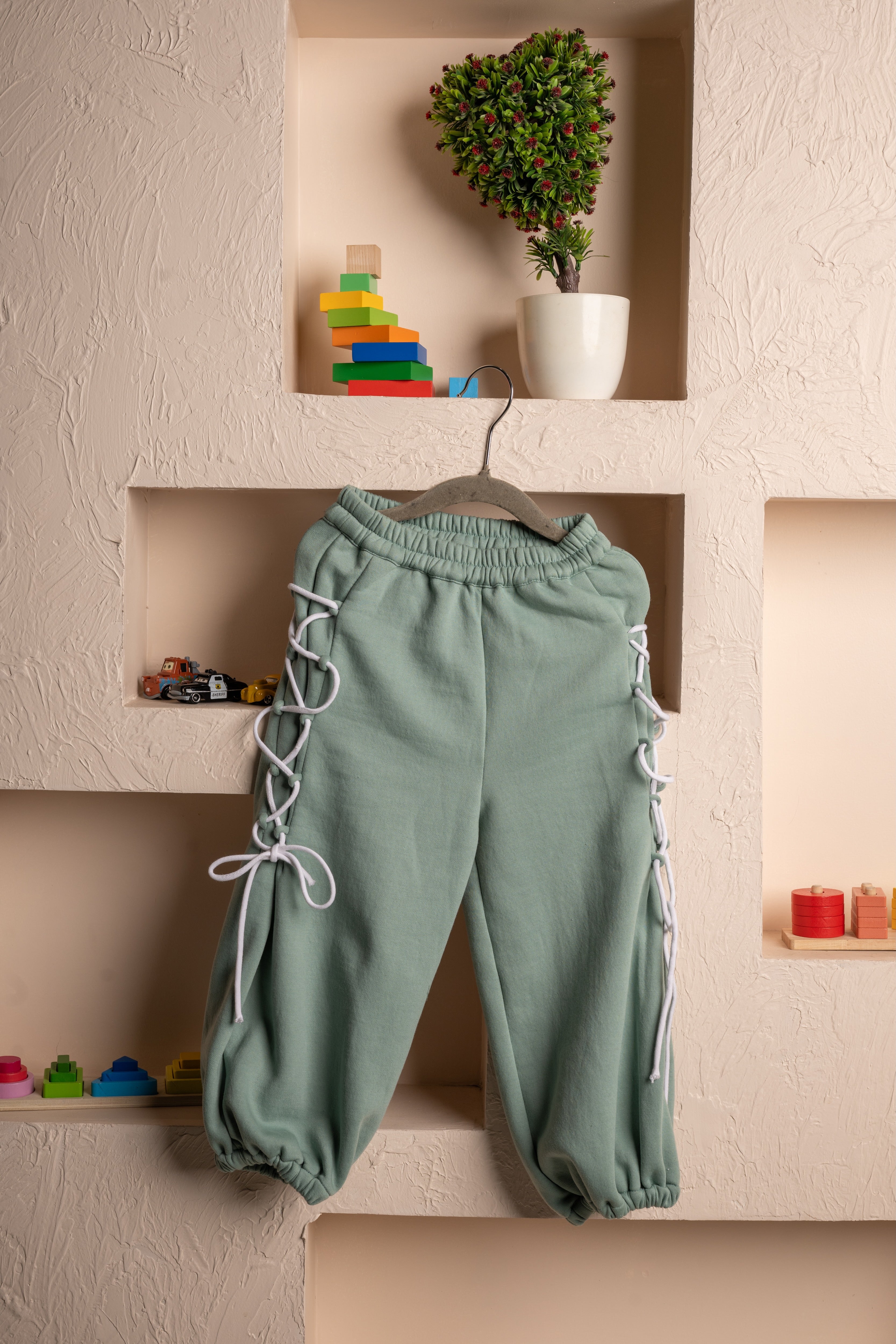 Elegant sage green jogger pants with white lace-up sides displayed on a textured beige wall niche