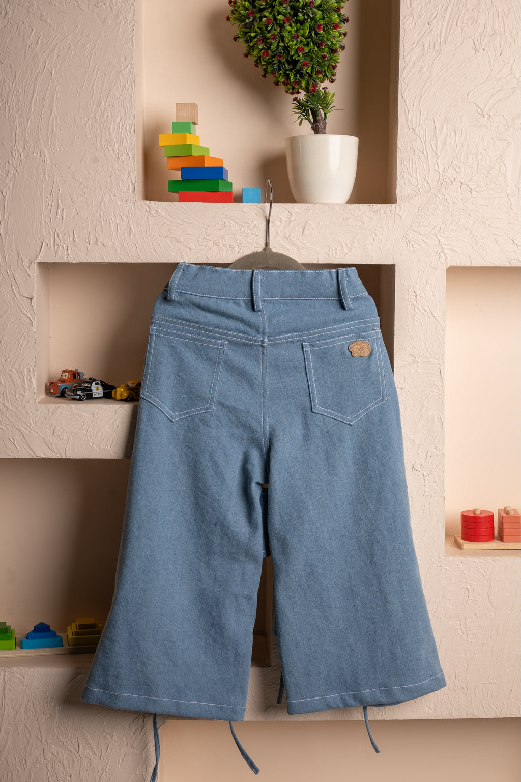 Elegant kid's wide-leg denim pants hanging on beige textured wall with colorful wooden toys and potted plant