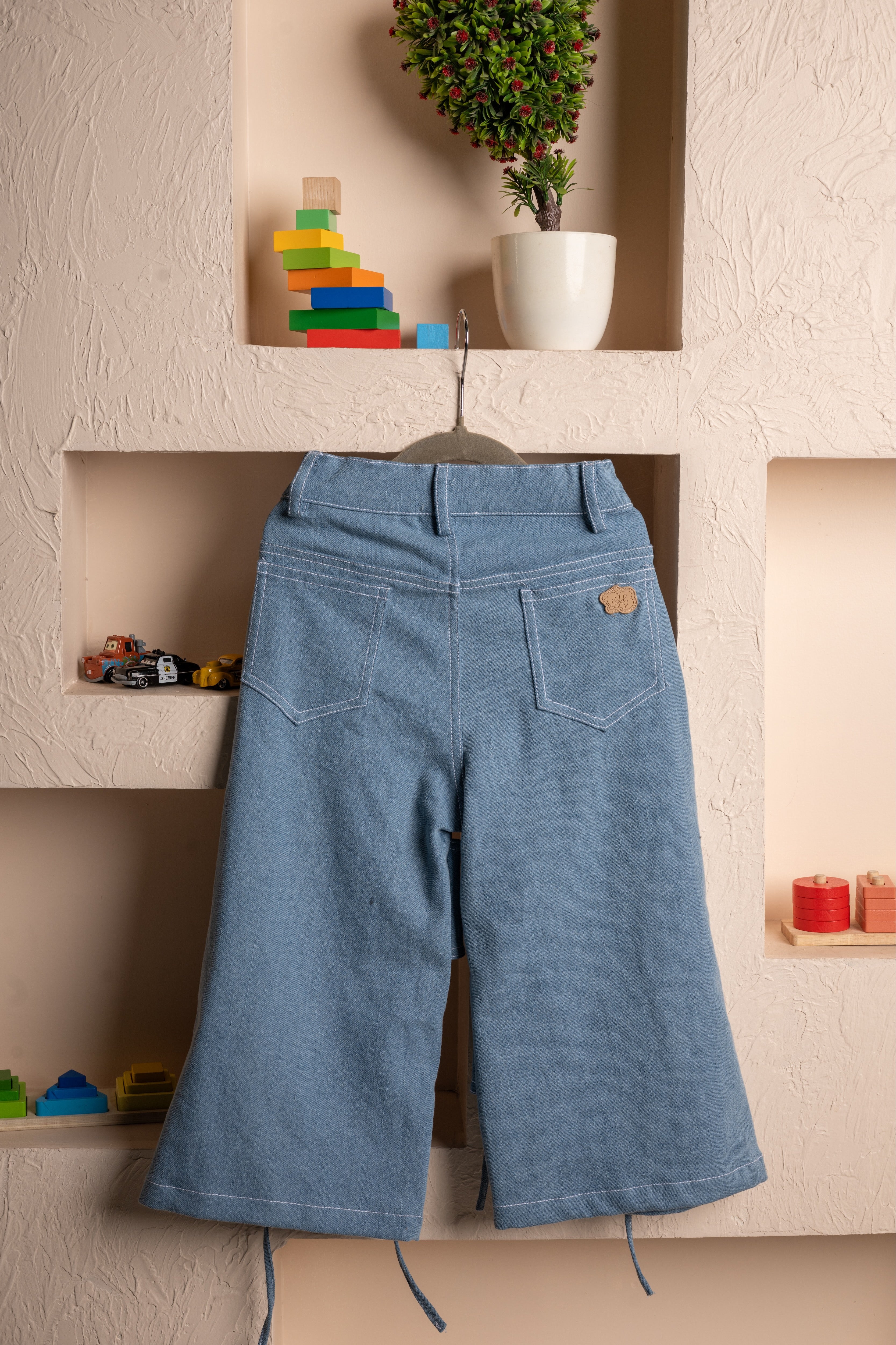Elegant kid's wide-leg denim pants hanging on beige textured wall with colorful wooden toys and potted plant